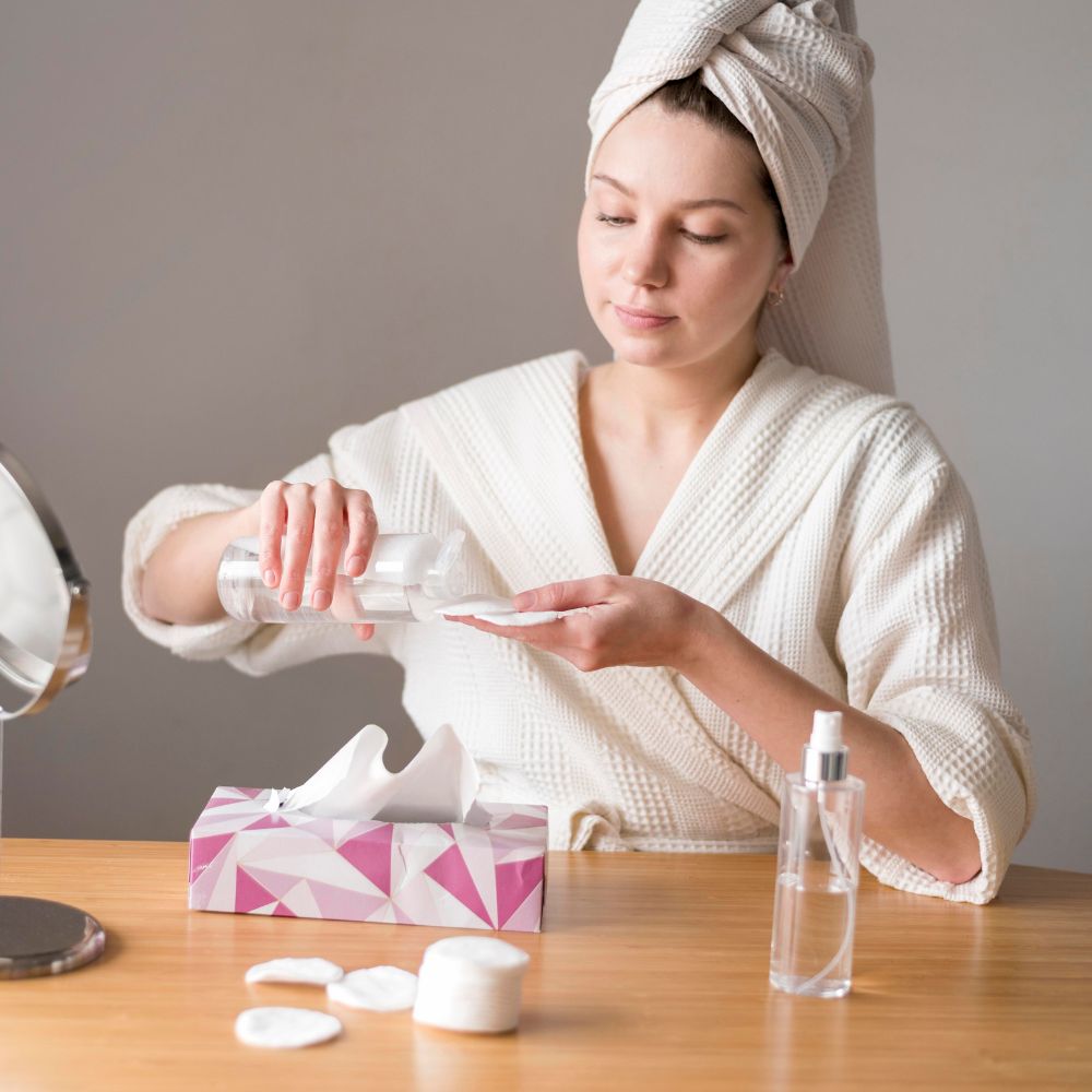 Woman in a robe applying cream to her face with a towel on her head, sitting at a table with skincare products.